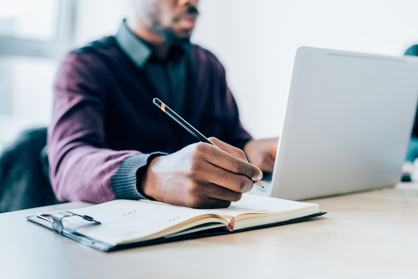 Person writing in notebook while working on laptop in office